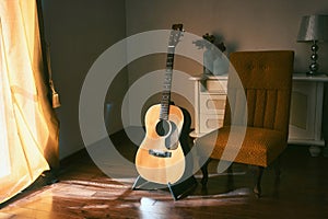 An acoustic Spanish guitar on a stand next to a chair in the moody shadows of a dark room