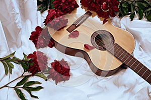 An acoustic wooden guitar lies on a white sheet surrounded by red peonies