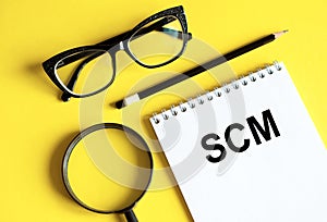 Acronym SCM supply chain management in notebook with pencil, magnifier, and glasses on a yellow background