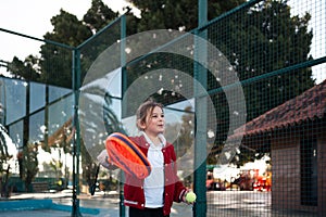 An action shot of a child reaching high to serve a padel tennis ball on an outdoor court, with palm trees in the