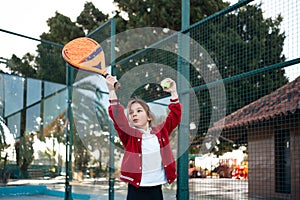 An action shot of a child reaching high to serve a padel tennis ball on an outdoor court, with palm trees in the