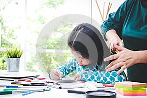 A cute little Asian girl is drawing and coloring, and the mother does her hair.