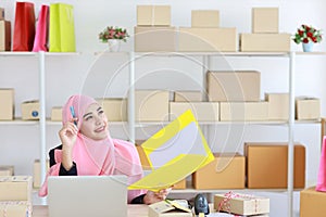 Active asian muslim woman wearing suit sitting and holding folder with computer and online package box delivery. Startup small