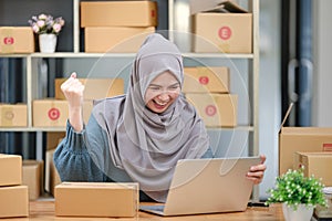 Active asian woman in blue muslim suit sitting and working with computer and online package box delivery. Startup small