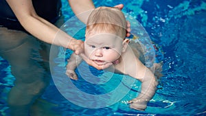 Active baby first swimming in woman`s hands in blue swimming pool. Cropped female hands holding carefully baby`s head