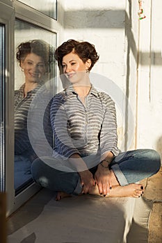 An active, beautiful middle-aged woman with a beautiful smile. Sitting on the windowsill near the window. Reflection in the window