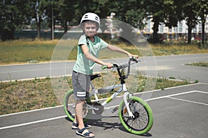 Active Caucasian child boy, in sports helmet, looking at camera, standing near his bicycle on the asphalt road outdoor.