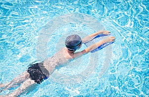 Active child (boy) in cap, sport goggles doing water sport with swim board in the swimming pool.