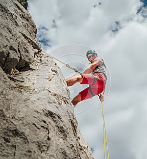 Active climber middle age man in protective helmet looking at camera while abseiling from cliff rock wall using rope with belay photo