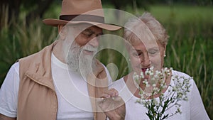 An active couple seniors is having fun in a meadow in a city park. An older lady holds a meadow flower in her hand