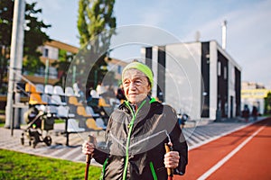 Active elderly Caucasian elderly women of 90 years practice Nordic walking with ski poles on a track with a red rubber coating.