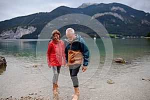 Active elderly couple hiking together in spring mountains, walking in cold water in mountain lake. Senior tourists