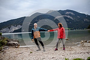 Active elderly couple hiking together in spring mountains, walking on pebble beach of mountain lake. Senior tourists