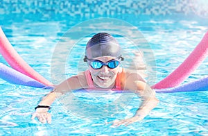 Active exercise senior woman (over age of 50) in goggles, cap doing water sport, aerobics with swim noodles.