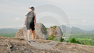 Active female hiker descends mountain trail with rich green landscape, Sigiriya Rock background. Solo traveler explores