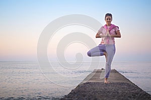 Active female practicing yoga vrikshasana tree pose hands namaste at sunset beach sea sky horizon