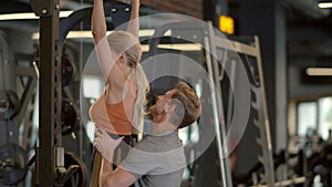 Active fit girl making pullups in fitness center. Trainer helping client at gym