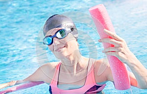 Active fitness senior woman (over age of 50) in goggles, cap doing water sport, aerobics with swim noodles.