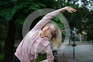 Active fitness woman in pink clothes and eyeglasses is stretching outdoor in the park during spring or summer morning