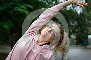 Active fitness woman in pink clothes and eyeglasses is stretching outdoor in the park during spring or summer morning