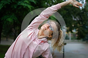 Active fitness woman in pink clothes and eyeglasses is stretching outdoor in the park during spring or summer morning