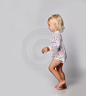 Active frolic blonde kid baby girl in jumpsuit one-piece with floral print pattern runs passes by camera. Side view.