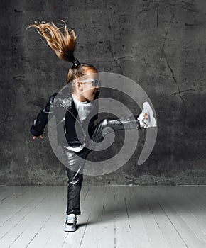 Active frolic kid girl in leather jacket, pants, shimmering sneakers and sunglasses is jumping lifting her leg high