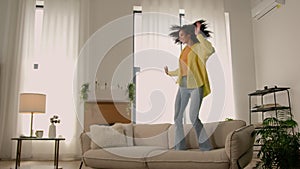 Active funny crazy African American girl young happy woman jumping on sofa couch in living room excited female jump