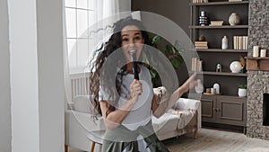 Active funny young woman looking at camera excitedly singing song holding tv remote as microphone having fun at home on