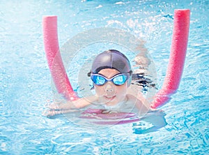 active, happy child in cap, sport goggles doing water sport with swim noodle in the swimming pool. Healthy lifestyle.