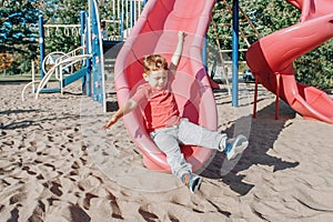 Active happy funny smiling Caucasian boy child sliding on playground schoolyard outdoors on summer sunny day. Kid having fun.