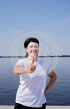 Active and happy senior woman in sportswear showing thumbs up working out near the riverside