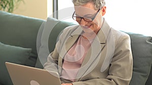 Active happy senior woman wearing glasses and short haircut sits on sofa and works remotely using laptop