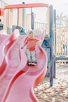 Active happy smiling Caucasian boy sliding on playground. Kid on schoolyard outdoors on summer sunny day. Child having fun outside