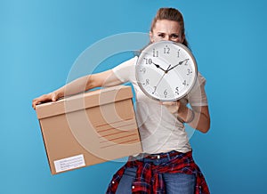 Active hipster with cardboard box hiding behind clock on blue