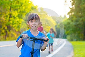 Active little girl riding a scooter on a park road on a summer day against her mother and sister in a blurred background. Family