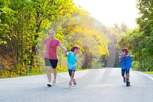 Active little girl riding a scooter on the street in the park on a summer day with her mother and sister jogging. Family spending