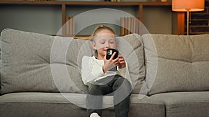 Active little kid girl jump on comfortable sofa in living room use modern gadget smartphone scroll pictures watch funny