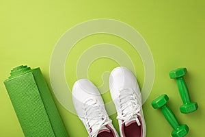 Active living concept. Top view photo of white sneakers green sports mat and dumbbells on isolated green background with copyspace