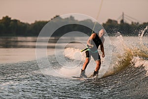 active man holds cable and energetically rides down on splashing wave on wakeboard.