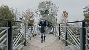 Active man jogging away on a metal bridge in a park with autumn trees. Athlete Doing Cardio Training. Jog Exercises