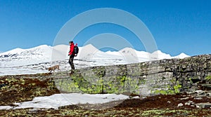 Active man in red jacket are walking in the wilderness with his dog with snow capped mountains in the back and blue sky
