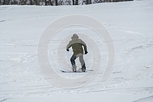 Active man snowboarder riding on slope. Man snowboarder snowboarding on white snow. Back view of male in khaki green coat and gray