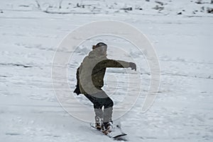 Active man snowboarder riding on slope. Man snowboarder snowboarding on white snow. Back view of male in khaki green coat and gray
