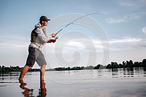 Active man is standing in shallow and fishing. He holds fly rod in hands. Man is twisting around reel to make spoon