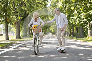 Active Mature Couple Riding Bike And Skateboard On Path In Park