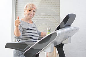 Active middle aged woman in sportswear showing thumb up on treadmill at home