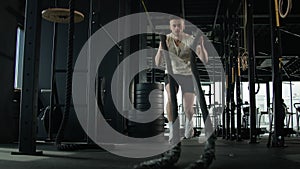 Active muscular athlete Caucasian sportsman man guy training in fitness gym workout squatting with heavy battle ropes