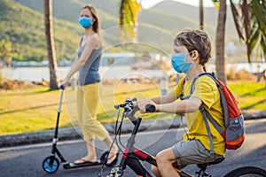 Active school kid boy and his mom in medical mask riding a bike with backpack on sunny day. Happy child biking on way to