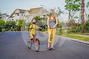 Active school kid boy and his mom in medical mask riding a bike with backpack on sunny day. Happy child biking on way to